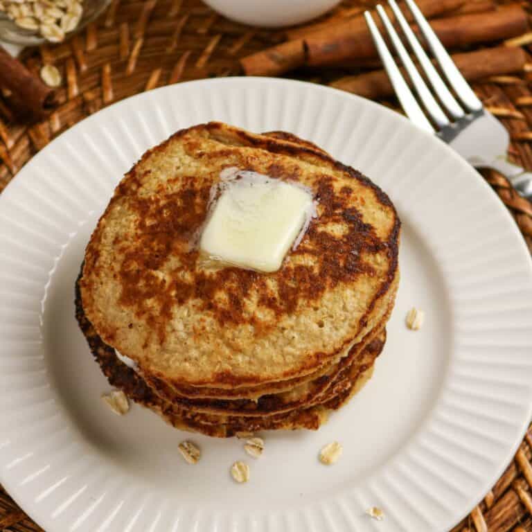 A stack of three pancakes sits on a white plate, topped with a pat of melting butter. A fork and cinnamon sticks are nearby, with some oats scattered on the plate and woven placemat background.