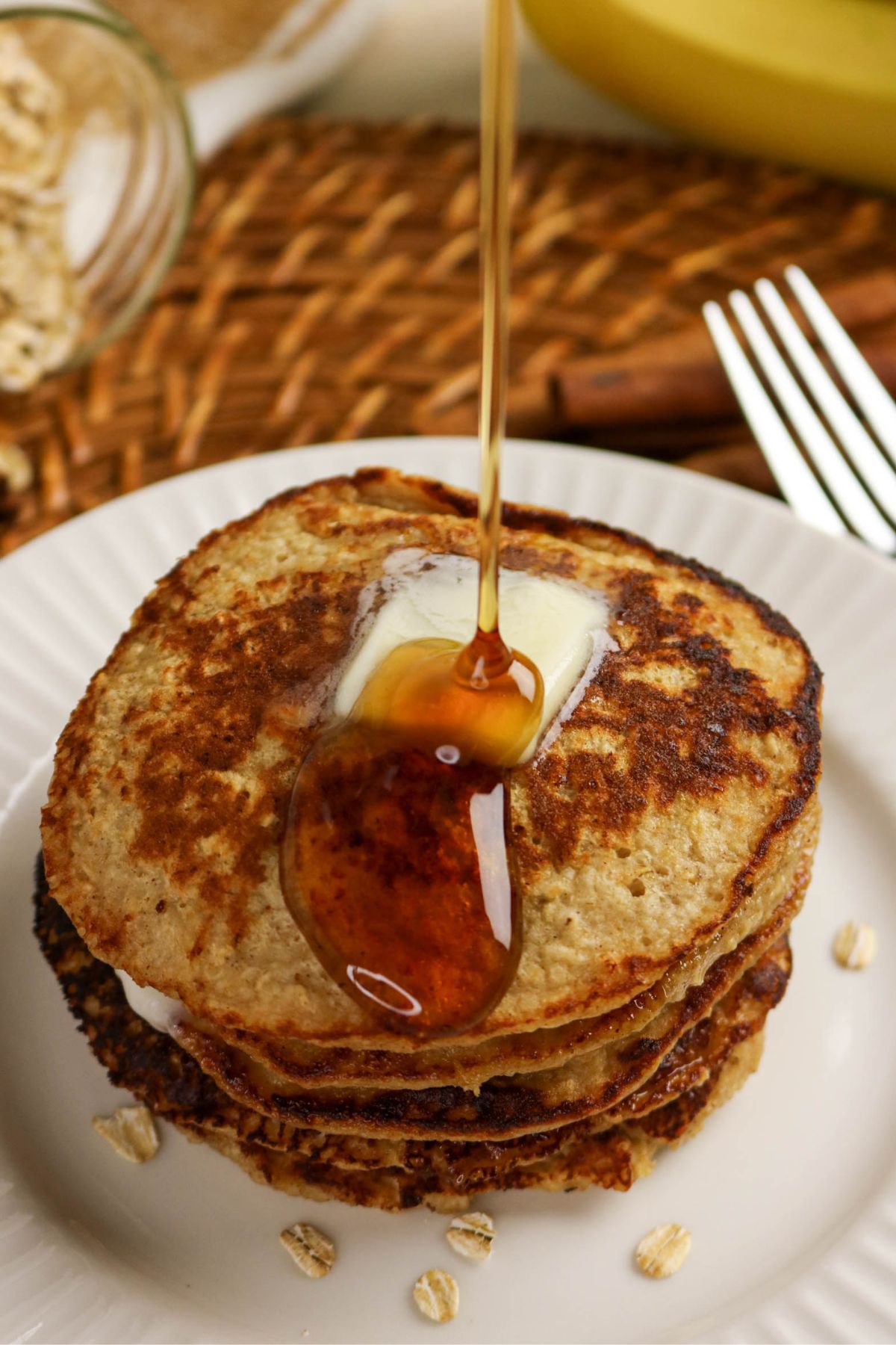 A stack of three pancakes topped with a pat of butter is shown on a white plate, with syrup being poured over the pancakes. A fork, knife, and oats are nearby on a woven placemat.