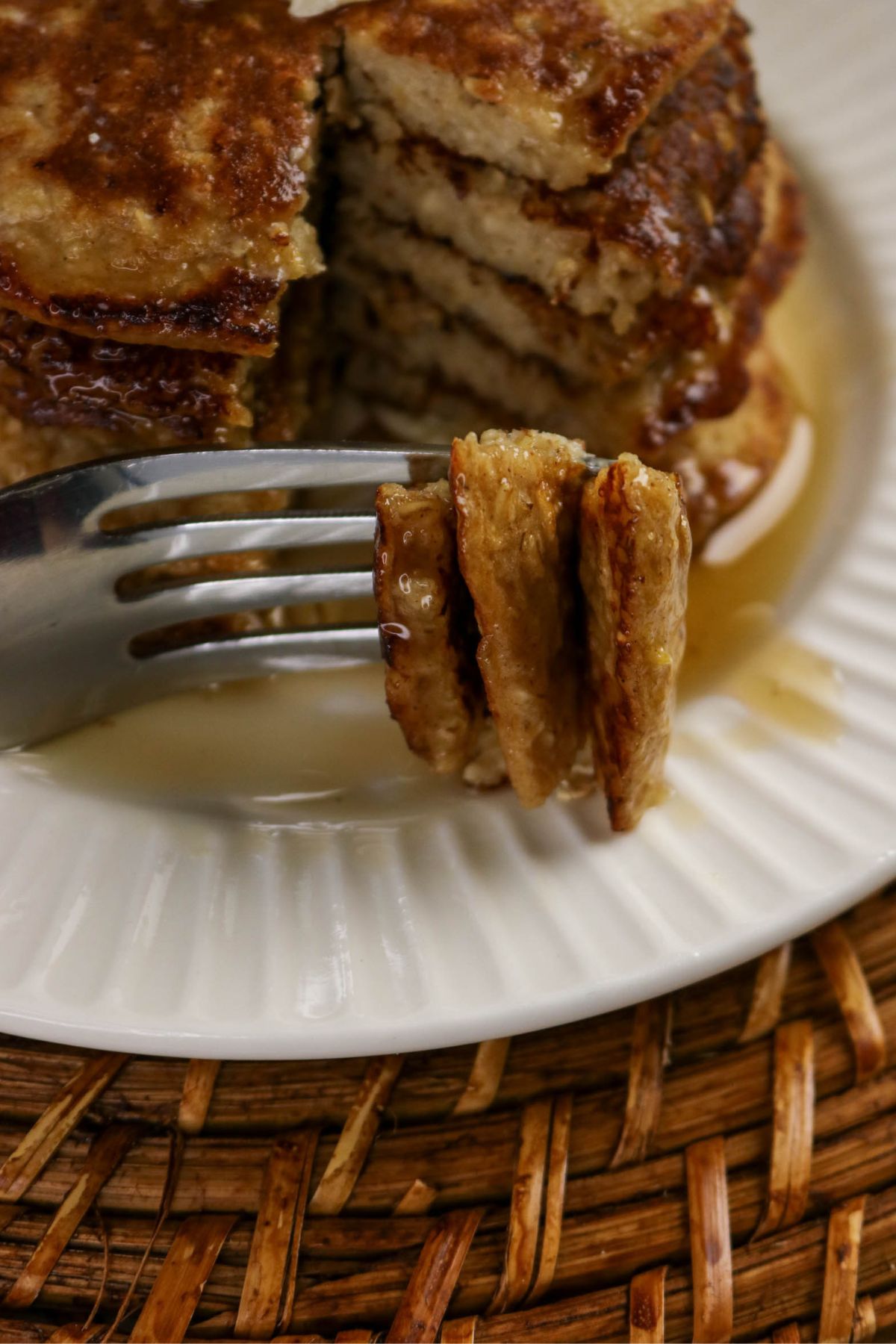 A close-up view of a stack of pancakes with syrup on a white plate. A fork in the foreground holds a bite-sized piece of pancake, and part of the stack is missing where it was cut.
