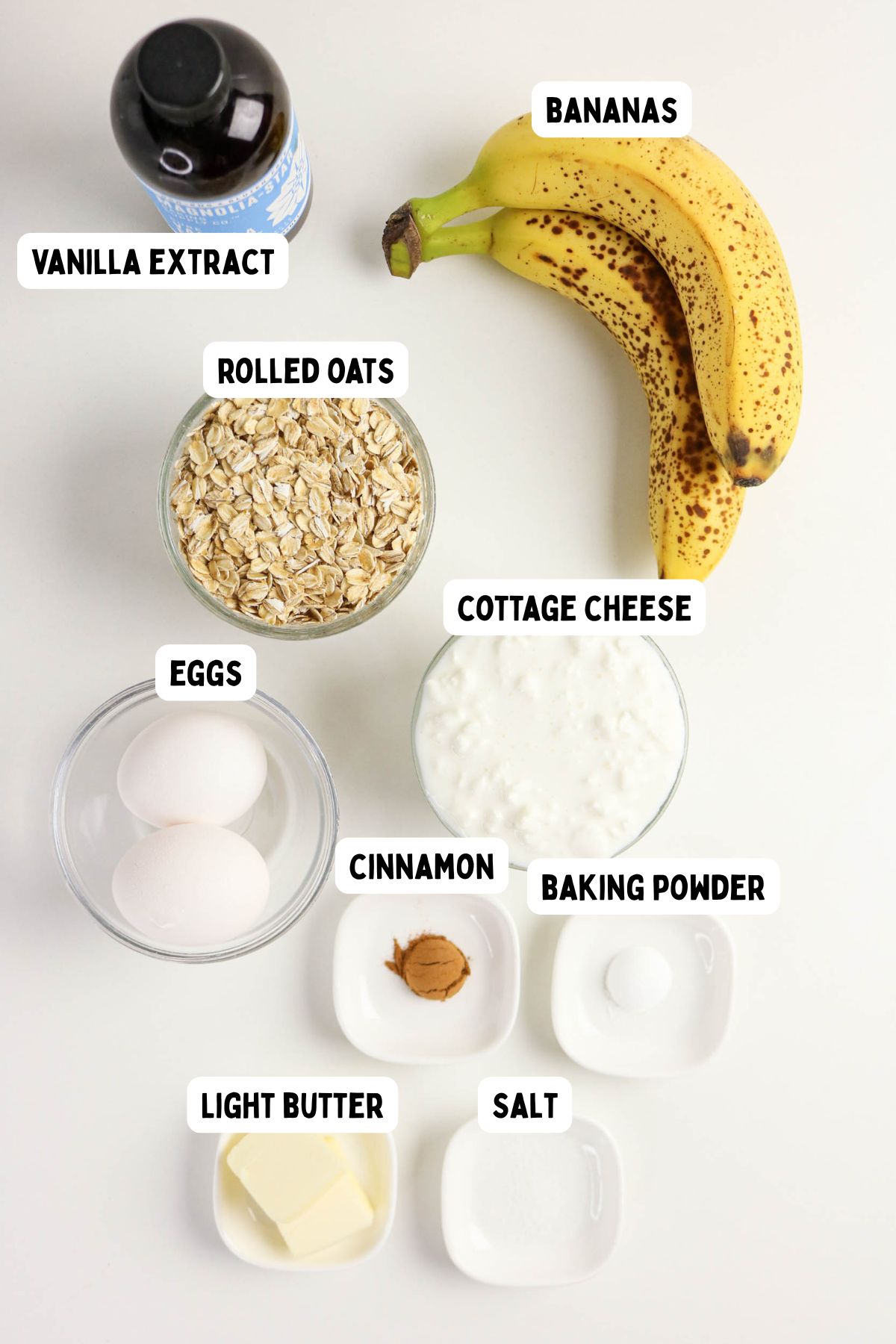 A top-down view of labeled baking ingredients: vanilla extract, bananas, rolled oats, cottage cheese, eggs, cinnamon, baking powder, light butter, and salt, each in separate containers on a white surface.
