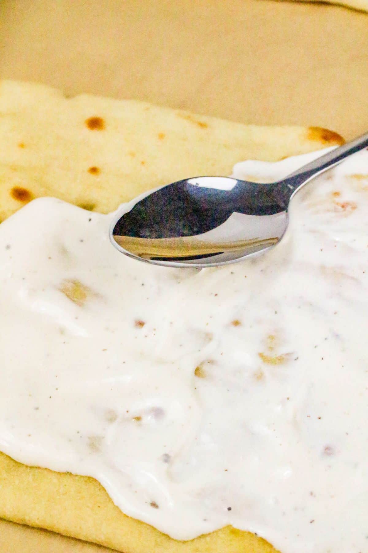A spoon spreads a thick, white sauce with small chunks onto dough, preparing the base for a delicious chicken bacon ranch pizza.