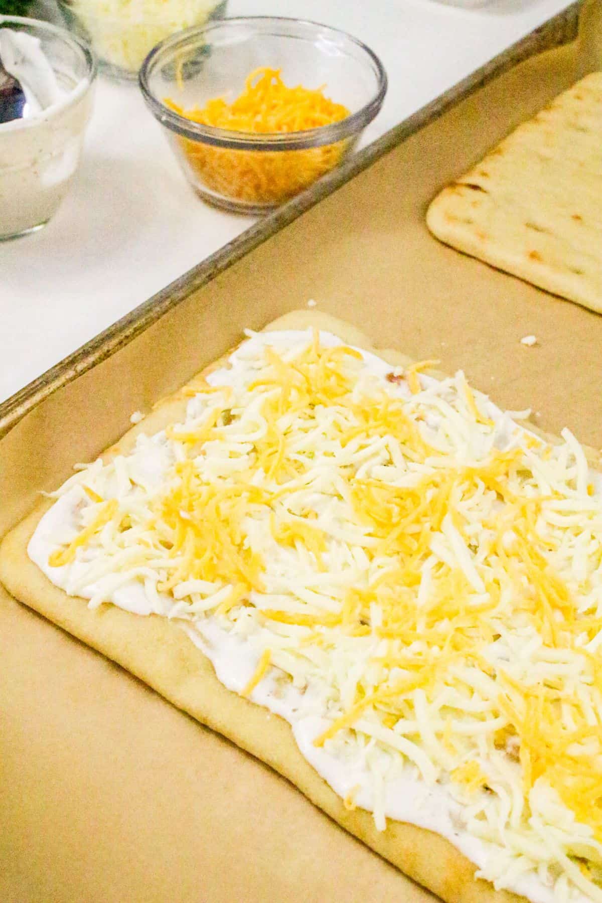 A rectangular piece of flatbread on a baking sheet is covered with a layer of ranch sauce and topped with shredded cheese, perfect for making chicken bacon ranch pizza. Small bowls of extra cheese and other toppings sit in the background.