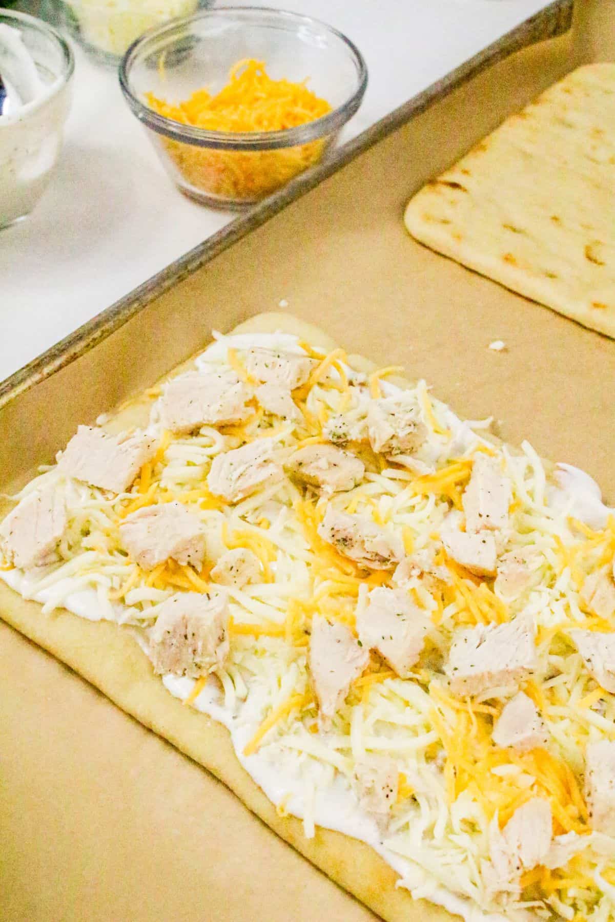 A flatbread on a baking sheet is topped with shredded cheese, small pieces of chicken, and creamy ranch sauce for a delicious chicken bacon ranch pizza. In the background, glass bowls hold extra cheese and other fresh ingredients.