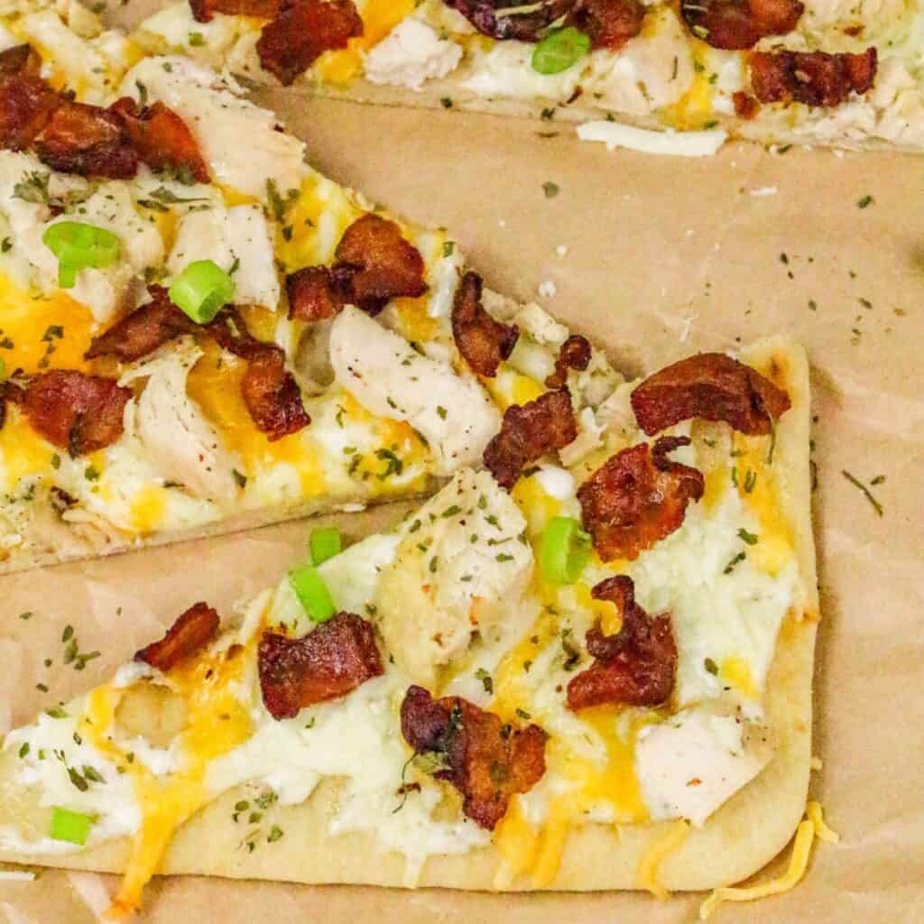 A close-up of two slices of chicken bacon ranch pizza on parchment paper, topped with pieces of chicken, bacon, melted cheese, green onion, and sprinkled herbs.