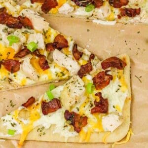 A close-up of two slices of chicken bacon ranch pizza on parchment paper, topped with pieces of chicken, bacon, melted cheese, green onion, and sprinkled herbs.