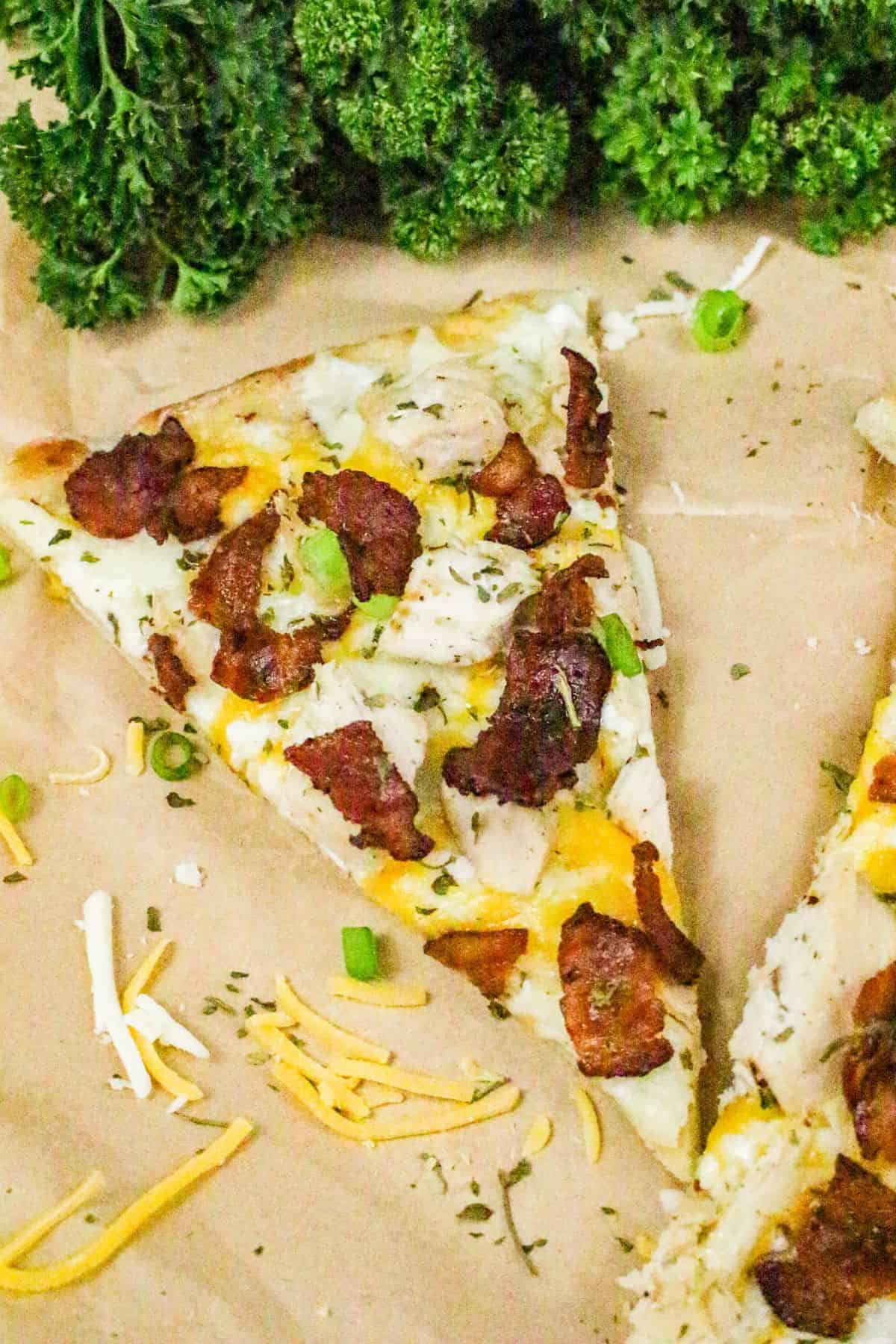 A slice of chicken bacon ranch pizza with pieces of bacon, chicken, green onions, and shredded cheese on a parchment surface, with fresh curly parsley in the background.