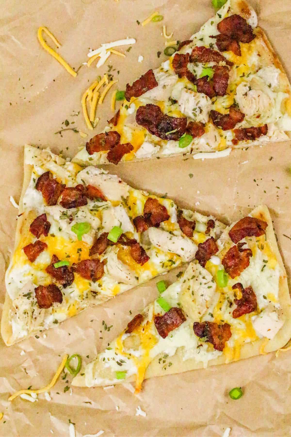 Three triangular slices of chicken bacon ranch pizza, topped with pieces of bacon, chicken, melted cheese, green onions, and herbs, are arranged on brown parchment paper with scattered cheese and green onion pieces.