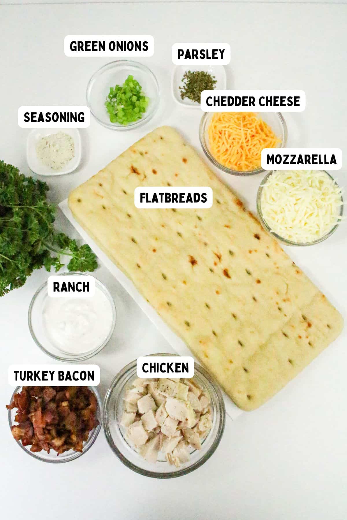 A flatbread on a white surface surrounded by labeled bowls of green onions, parsley, seasoning, cheddar cheese, mozzarella, ranch, turkey bacon, and diced chicken—perfect ingredients for a delicious chicken bacon ranch pizza.