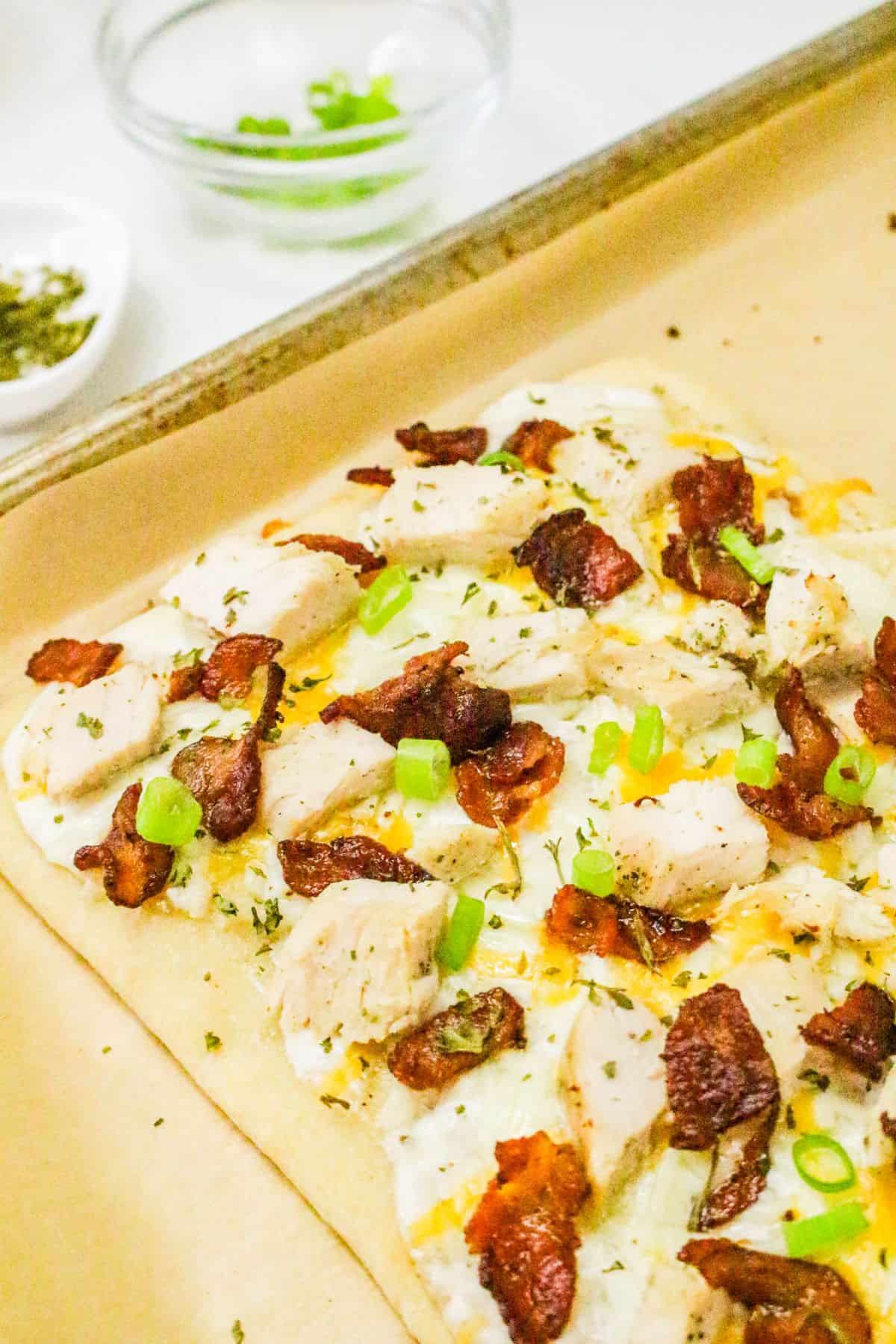 A chicken bacon ranch pizza flatbread topped with cooked chicken, crispy bacon, melted cheese, and sliced green onions sits on parchment paper on a baking sheet. Small bowls with extra toppings are in the background.