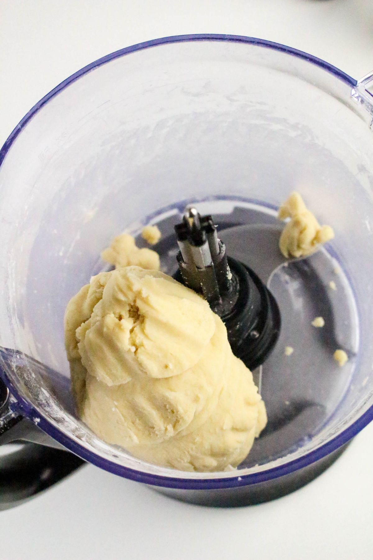 A close-up of a food processor bowl containing a ball of cookie dough, with some dough bits stuck to the sides and blades visible in the center.