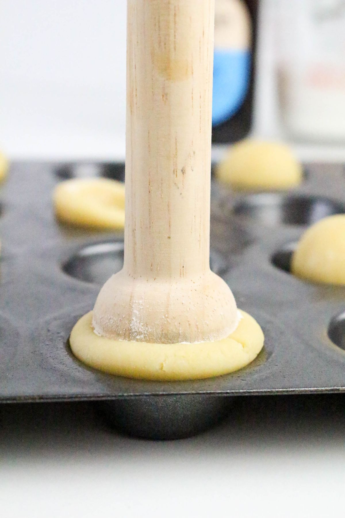 A wooden tool is pressing into a ball of dough on a baking tray, shaping it inside a round metal mold. More dough balls and baking ingredients are visible in the background.