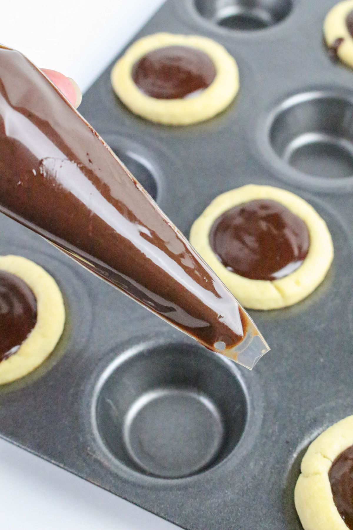 A hand holds a piping bag filled with chocolate ganache, positioned above a muffin tin containing several baked cookies with chocolate filling.