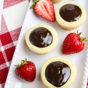 A white plate with three round cookies topped with glossy chocolate filling, accompanied by three fresh strawberries, one of which is sliced in half. The plate is placed on a red and white checkered cloth.