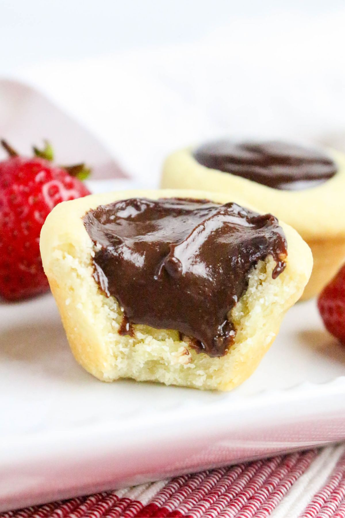 A close-up of a tart with a bite taken out, revealing a creamy chocolate filling. The tart is on a white plate, with whole strawberries and another tart in the background.