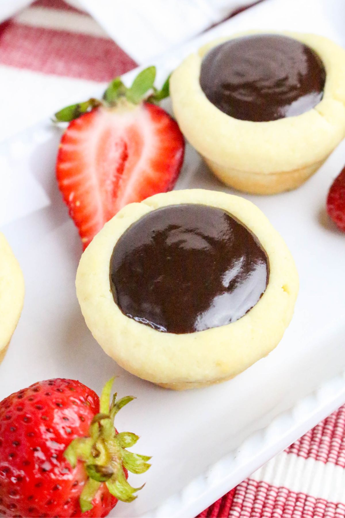 A close-up of two round cookies filled with glossy chocolate ganache on a white plate, with fresh strawberries and a sliced strawberry beside them.