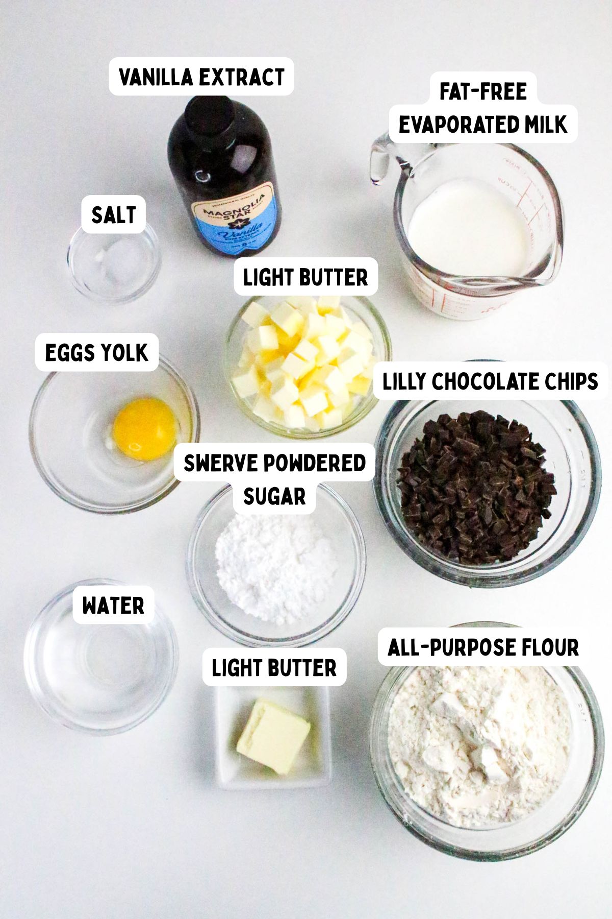 A top-down view of baking ingredients in bowls and measuring cups, labeled as vanilla extract, fat-free evaporated milk, salt, eggs yolk, light butter, Swerve powdered sugar, Lily chocolate chips, water, and all-purpose flour.