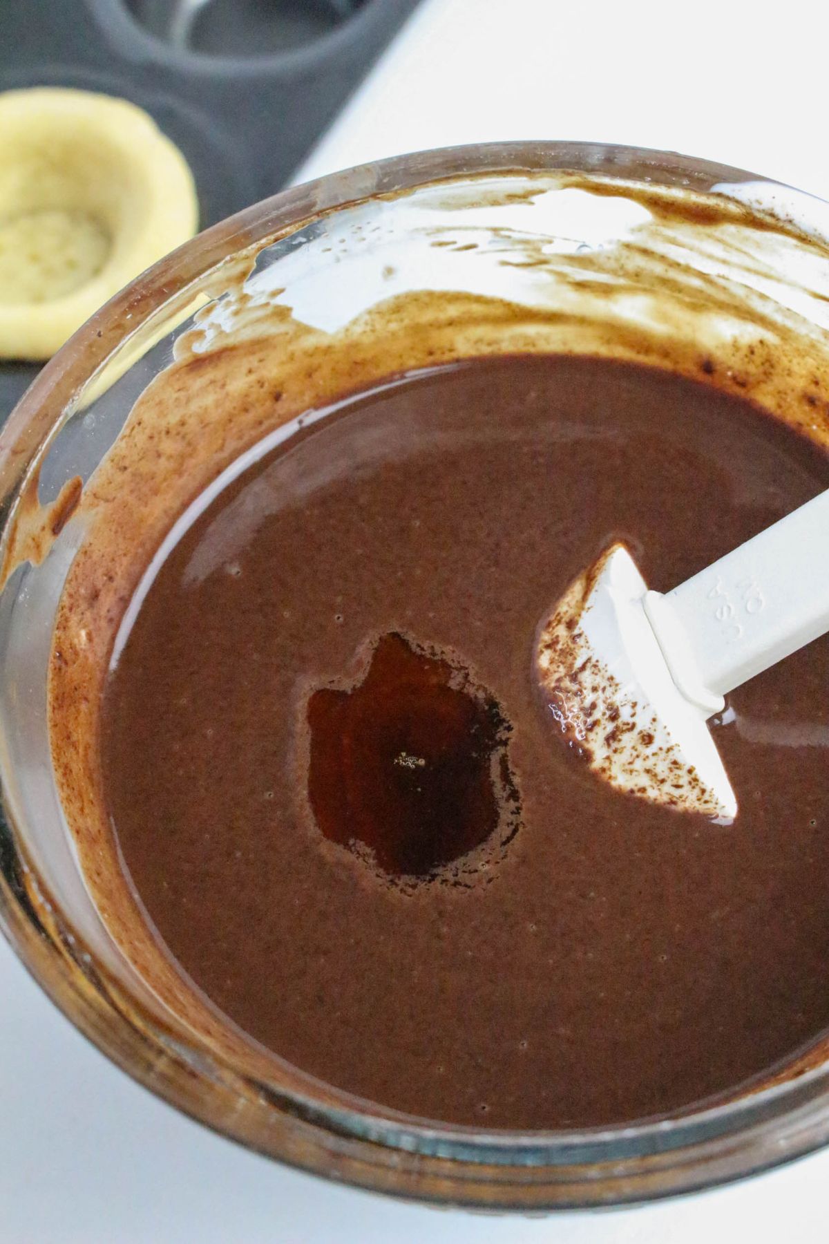 A glass bowl filled with chocolate batter, with a white spatula resting inside and some batter clinging to the sides of the bowl. A partially visible unbaked tart shell is in the background.