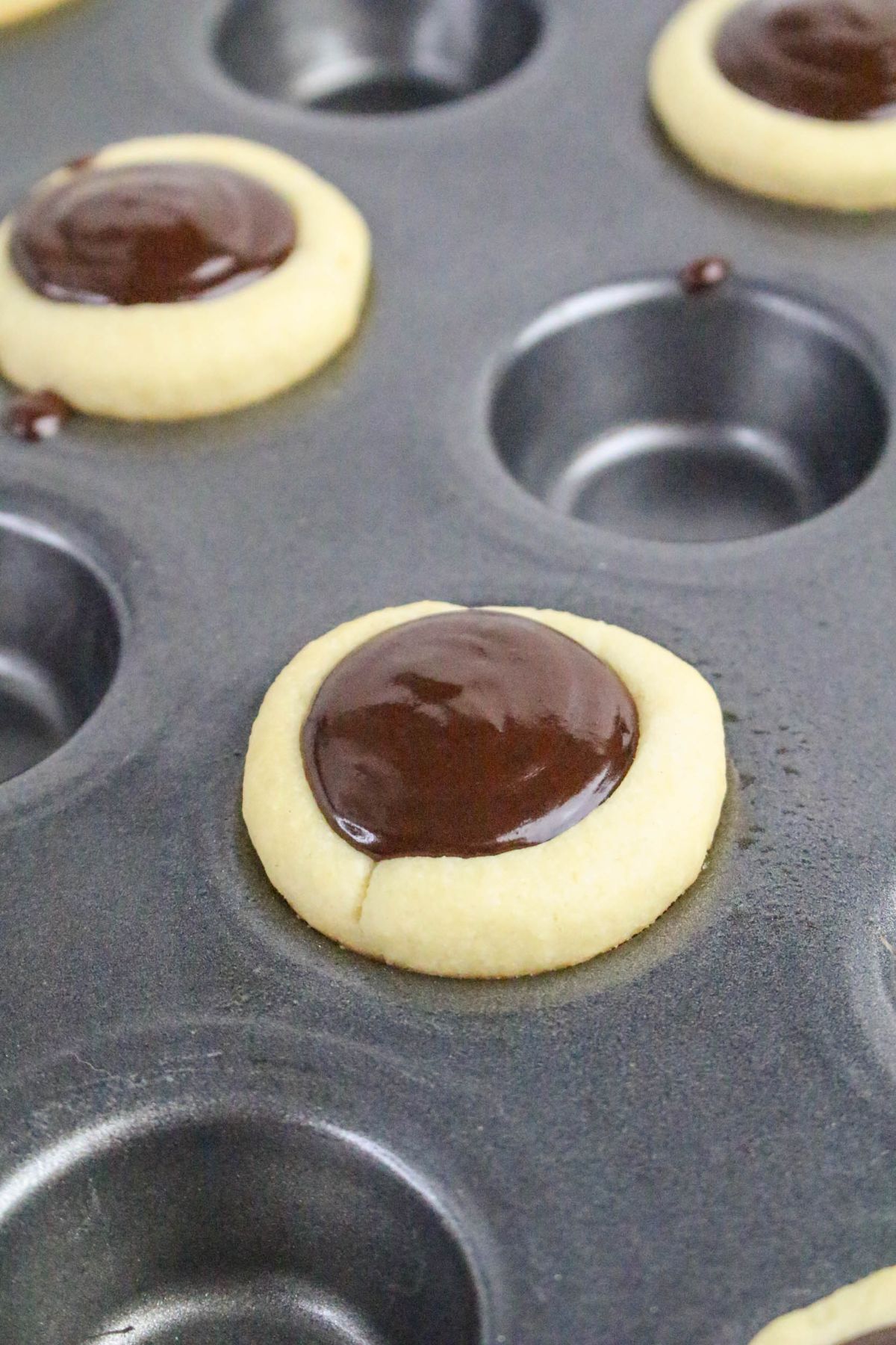 A close-up of a muffin tin holding thumbprint cookies with a glossy chocolate filling in the center, some mold cavities empty, and bits of chocolate visible on the pan.