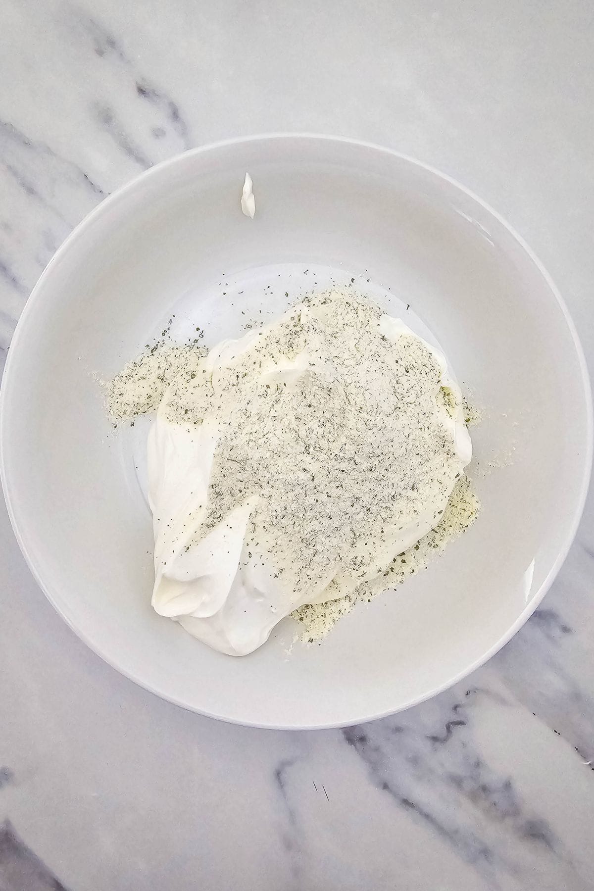 A white bowl on a marble surface contains a mound of white creamy substance topped with a generous amount of dry, speckled seasoning mix.
