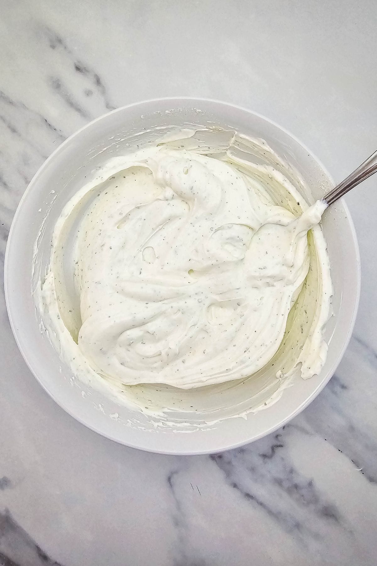 A white bowl filled with thick, creamy mixture sits on a marble surface. A spoon is resting in the bowl, with the creamy mixture slightly swirled around it.