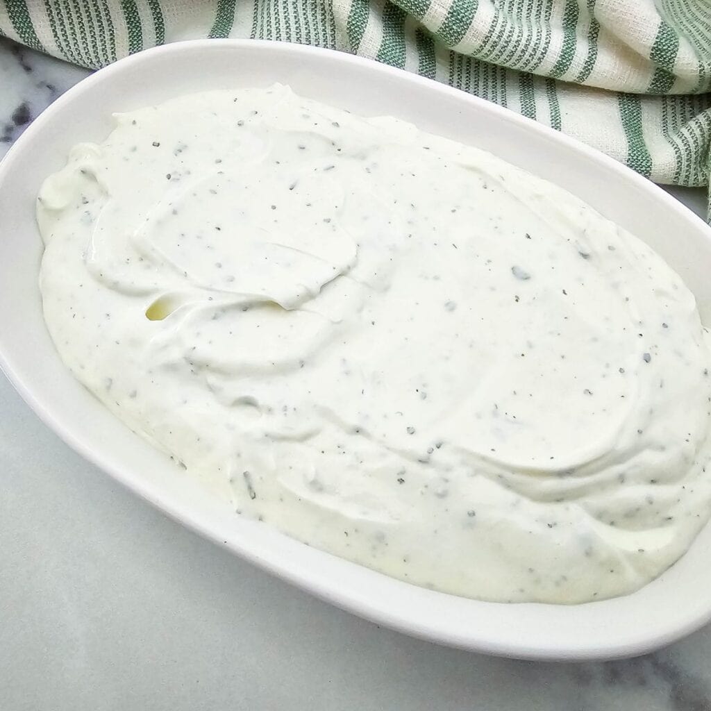 A white oval dish filled with creamy white ranch dressing speckled with herbs, placed on a light surface next to a green and white striped cloth.