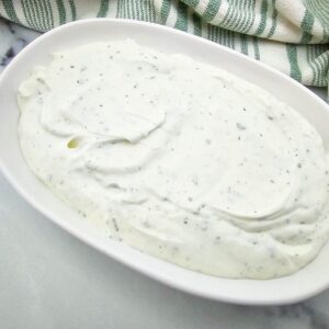 A white oval dish filled with creamy white ranch dressing speckled with herbs, placed on a light surface next to a green and white striped cloth.