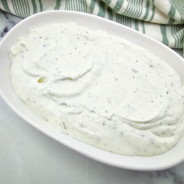A white oval dish filled with creamy white ranch dressing speckled with herbs, placed on a light surface next to a green and white striped cloth.