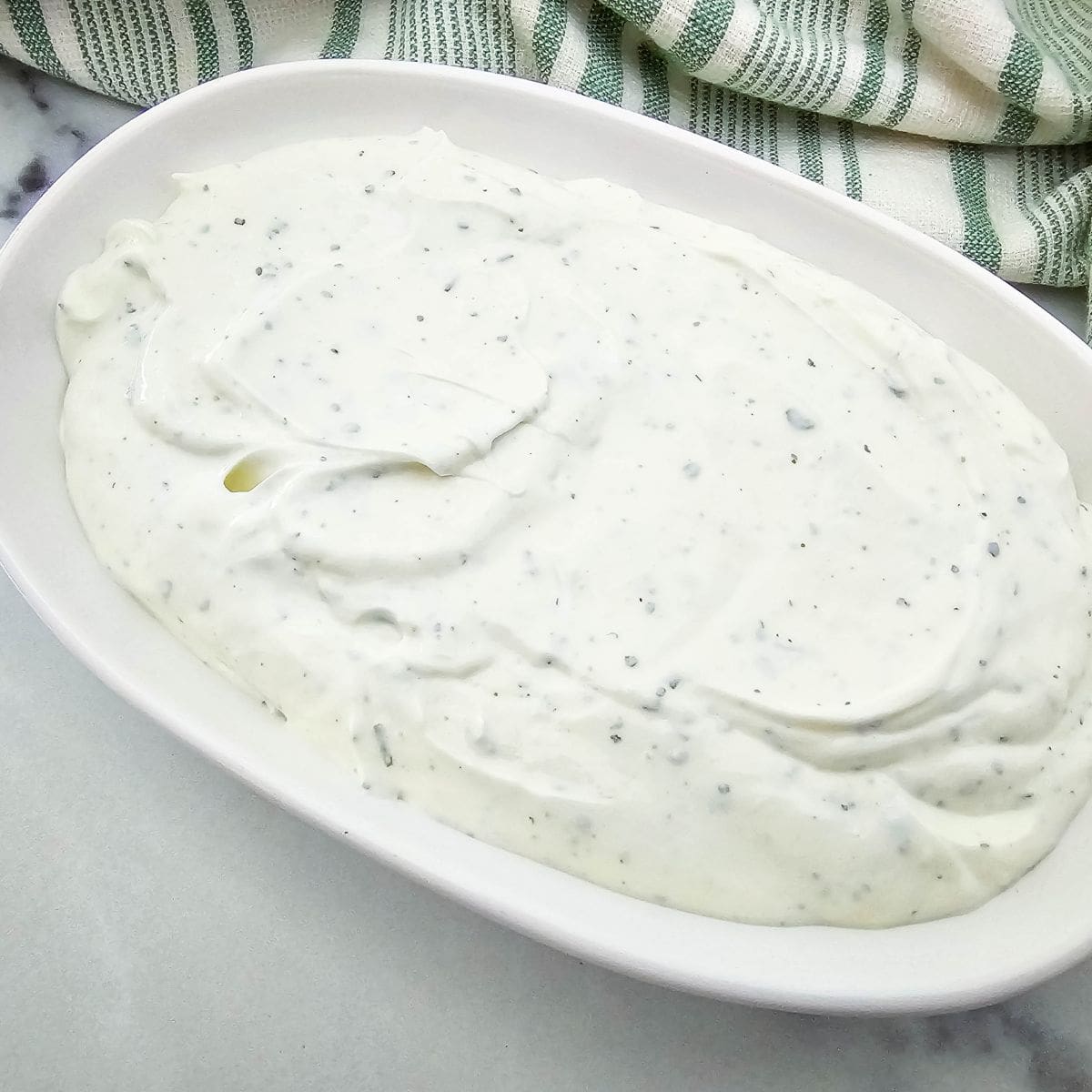 A white oval dish filled with creamy white ranch dressing speckled with herbs, placed on a light surface next to a green and white striped cloth.