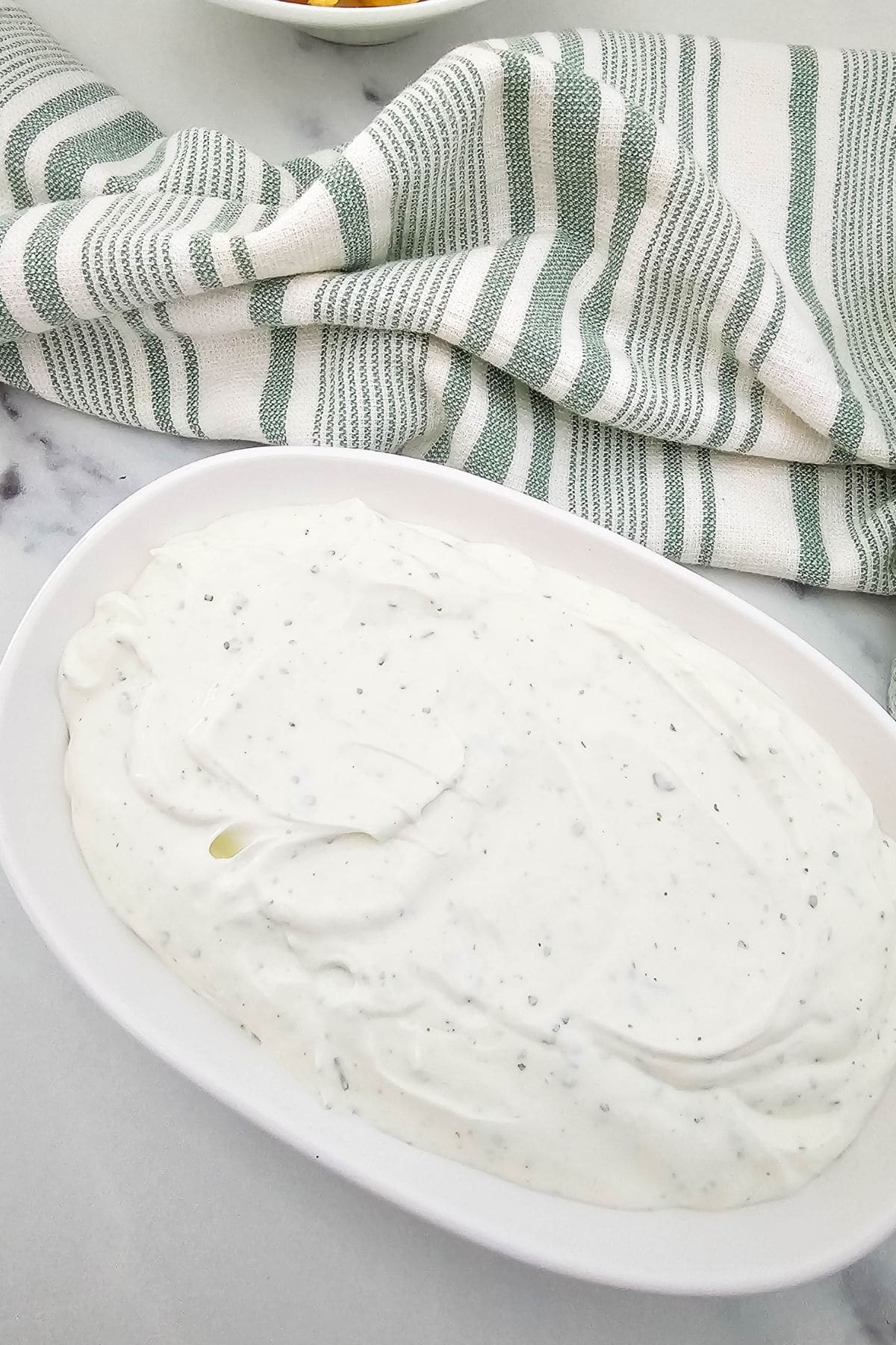 A white oval dish filled with a thick, white creamy sauce speckled with herbs, set on a light surface with a green and white striped towel in the background.