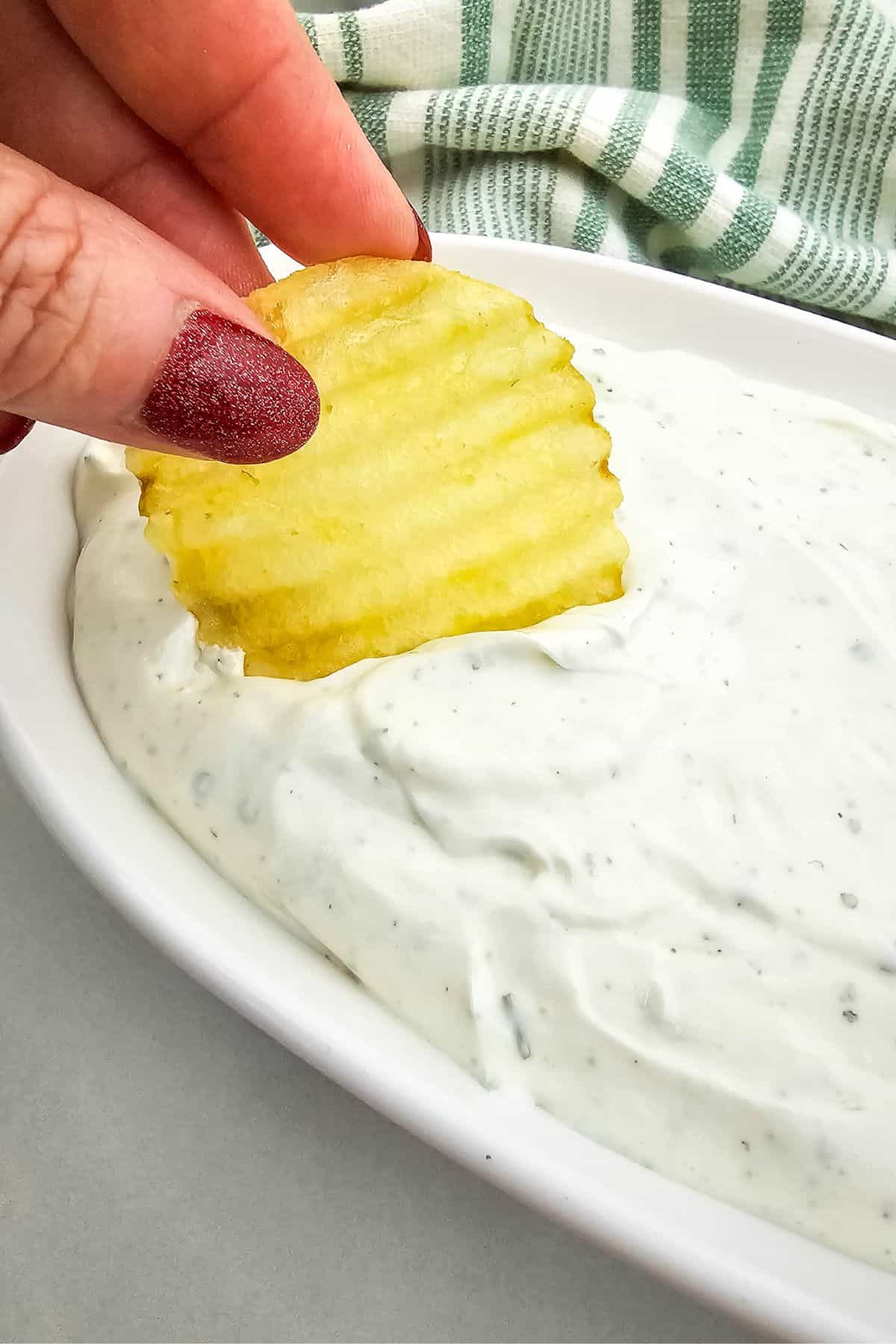 A hand with red nail polish dips a ridged potato chip into a white, creamy dip in a rectangular dish. A green and white striped cloth is visible in the background.