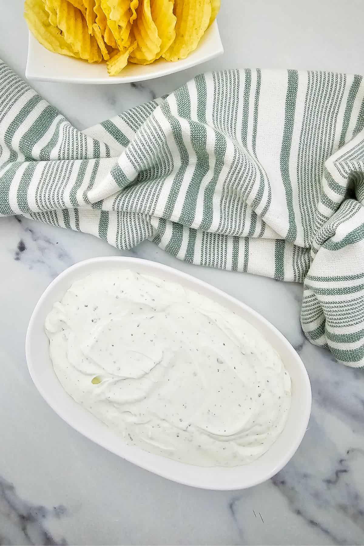 A white oval dish filled with creamy dip sits on a marble surface next to a green and white striped cloth. A plate of ridged potato chips is partially visible in the background.