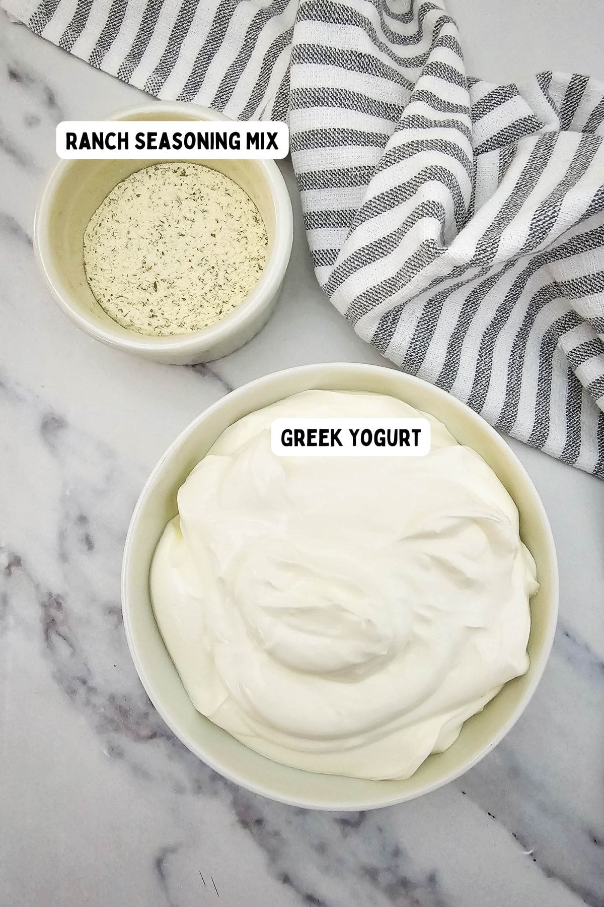 A bowl of ranch seasoning mix and a bowl of Greek yogurt on a marble surface, with a gray and white striped cloth near the top right corner. Each bowl is labeled accordingly.