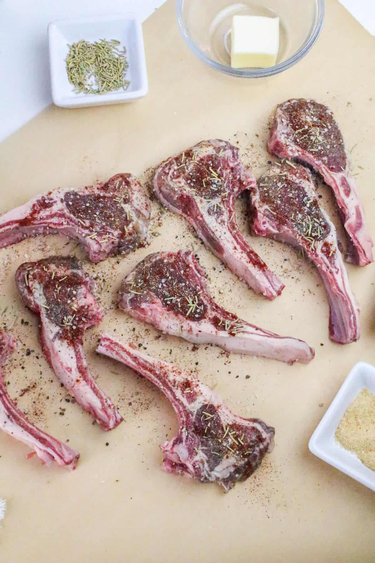 Six raw lamb chops are arranged on parchment paper, seasoned with herbs and spices, ready to be transformed into perfectly pan seared lamb chops. Surrounding the chops are small dishes containing dried herbs, a pat of butter, and breadcrumbs.