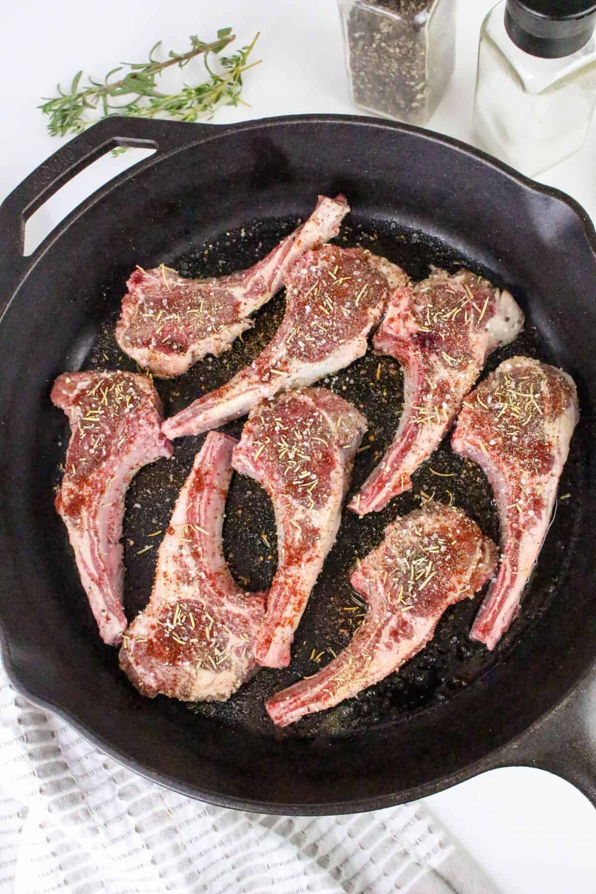 Six raw, seasoned lamb chops, perfect for pan seared lamb chops, are arranged in a black cast iron skillet. A sprig of herbs, a pepper grinder, and a salt shaker are visible nearby, along with a white cloth.