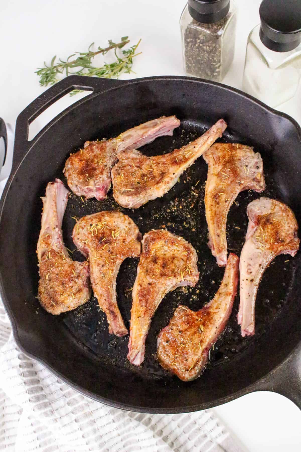 Seven seasoned pan seared lamb chops cooking in a black cast iron skillet. Black pepper and salt shakers, as well as a sprig of rosemary, are visible in the background. A white textured cloth is partially seen in the foreground.