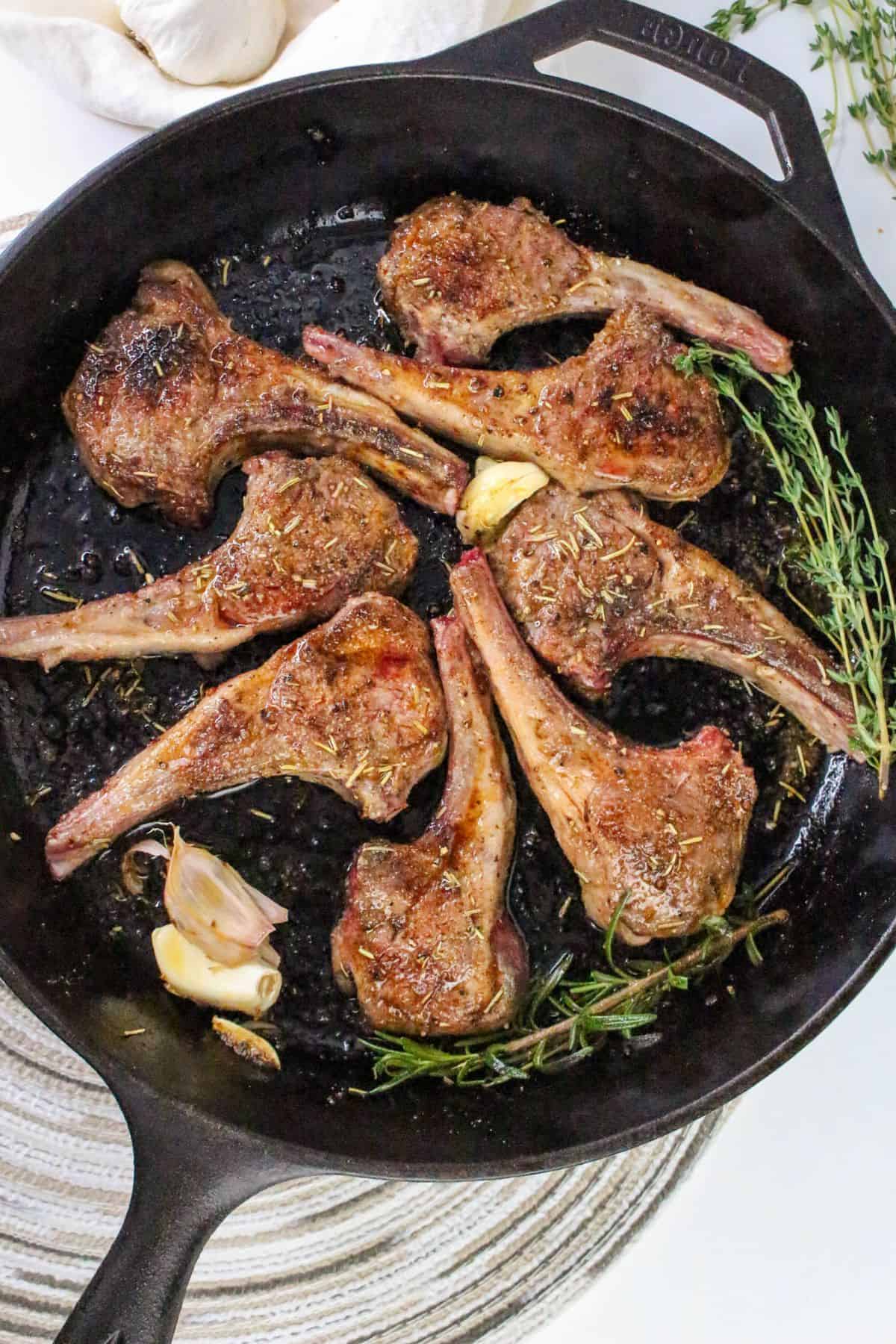 Six pan seared lamb chops in a cast iron skillet with sprigs of rosemary, thyme, and garlic cloves on a light woven mat. The meat appears browned and seasoned.