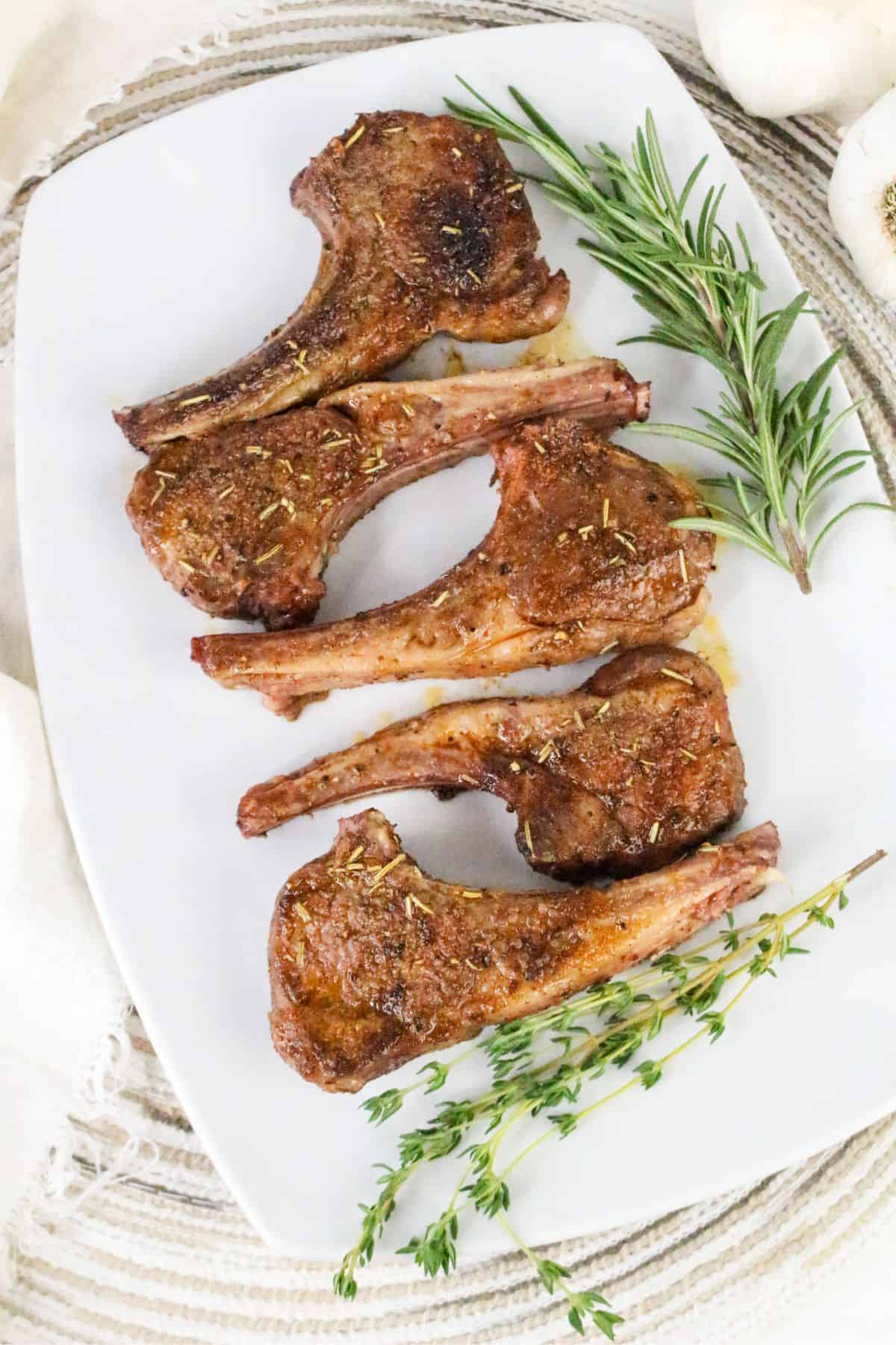 Five pan seared lamb chops are arranged on a white plate, garnished with fresh rosemary and thyme sprigs. The plate is set on a woven placemat with a head of garlic visible in the top right corner.