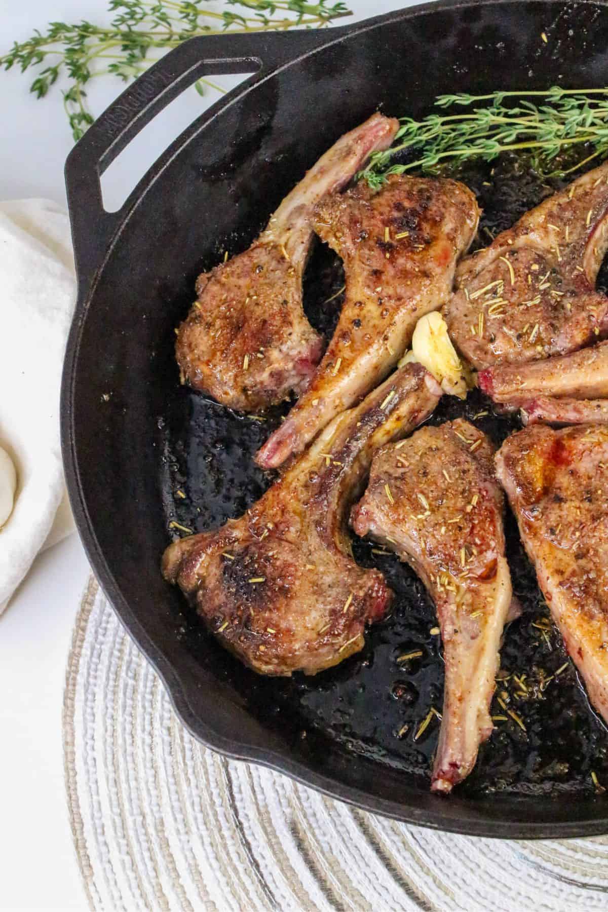 Five pan seared lamb chops with herbs and a garlic clove are arranged in a black cast iron skillet. A sprig of fresh rosemary is also present. The skillet sits on a round woven mat, with a white napkin nearby.