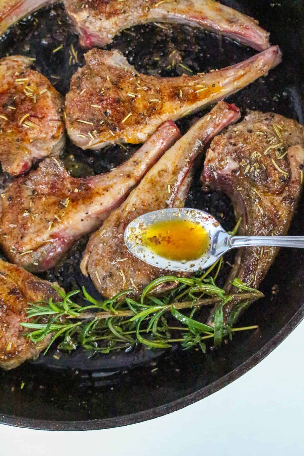 Five seasoned pan seared lamb chops cook in a dark pan with fresh rosemary. A metal spoon with brown drippings is held above the meat, suggesting basting. The surface appears caramelized and glistening with oil or butter.