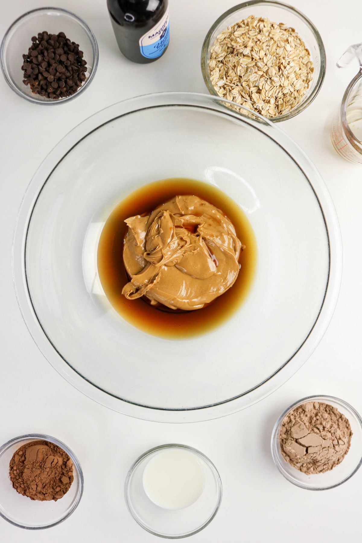 A glass bowl with peanut butter and liquid sits in the center, surrounded by small bowls of chocolate chips, rolled oats, cocoa powder, flour, milk, and a bottle of vanilla extract on a white surface.