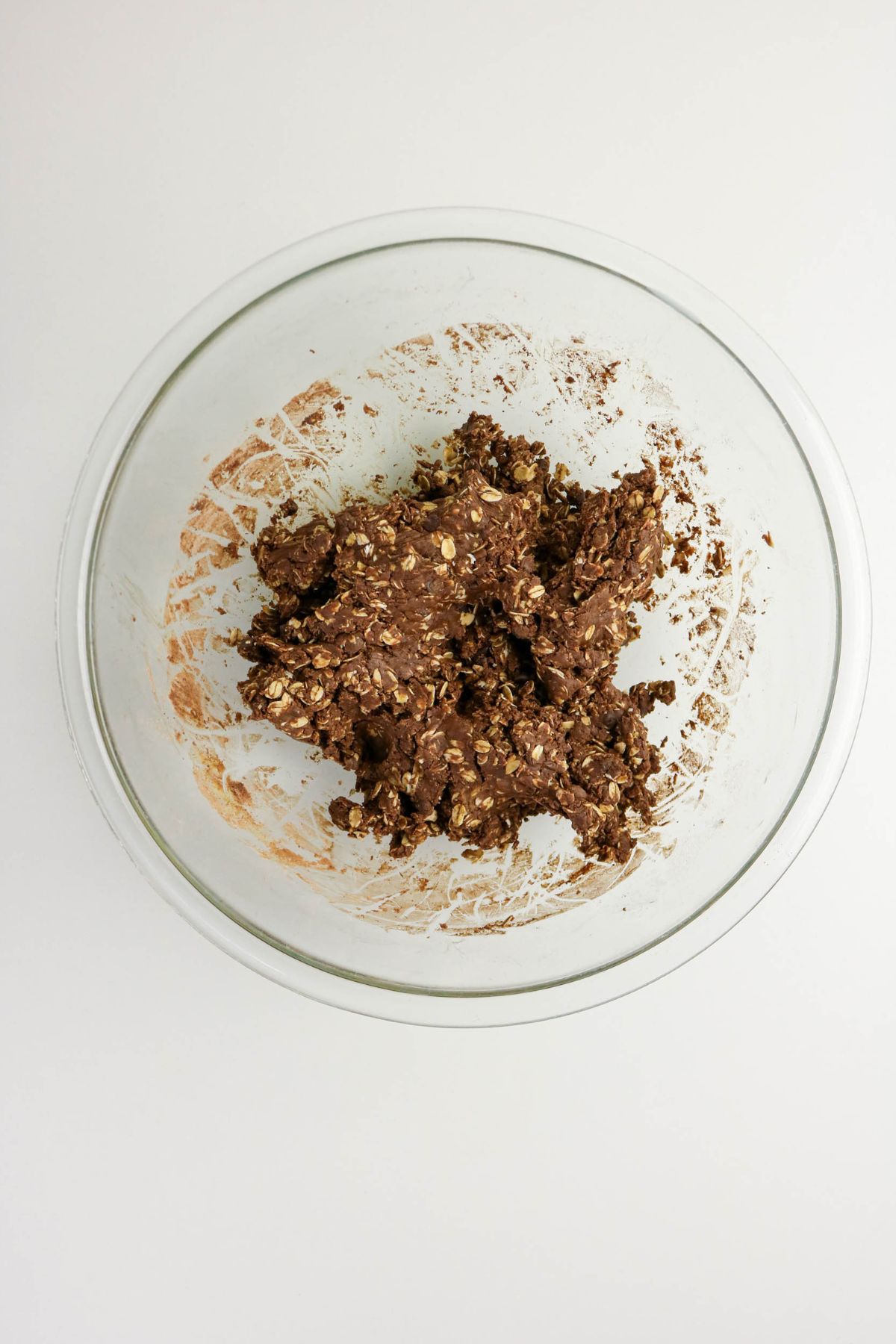 A clear glass mixing bowl containing chocolate oat cookie dough, partially mixed, sits on a white surface.