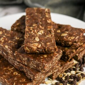 A close-up of several chocolate oat granola bars stacked on a white plate, with scattered oats and chocolate chips beside them.