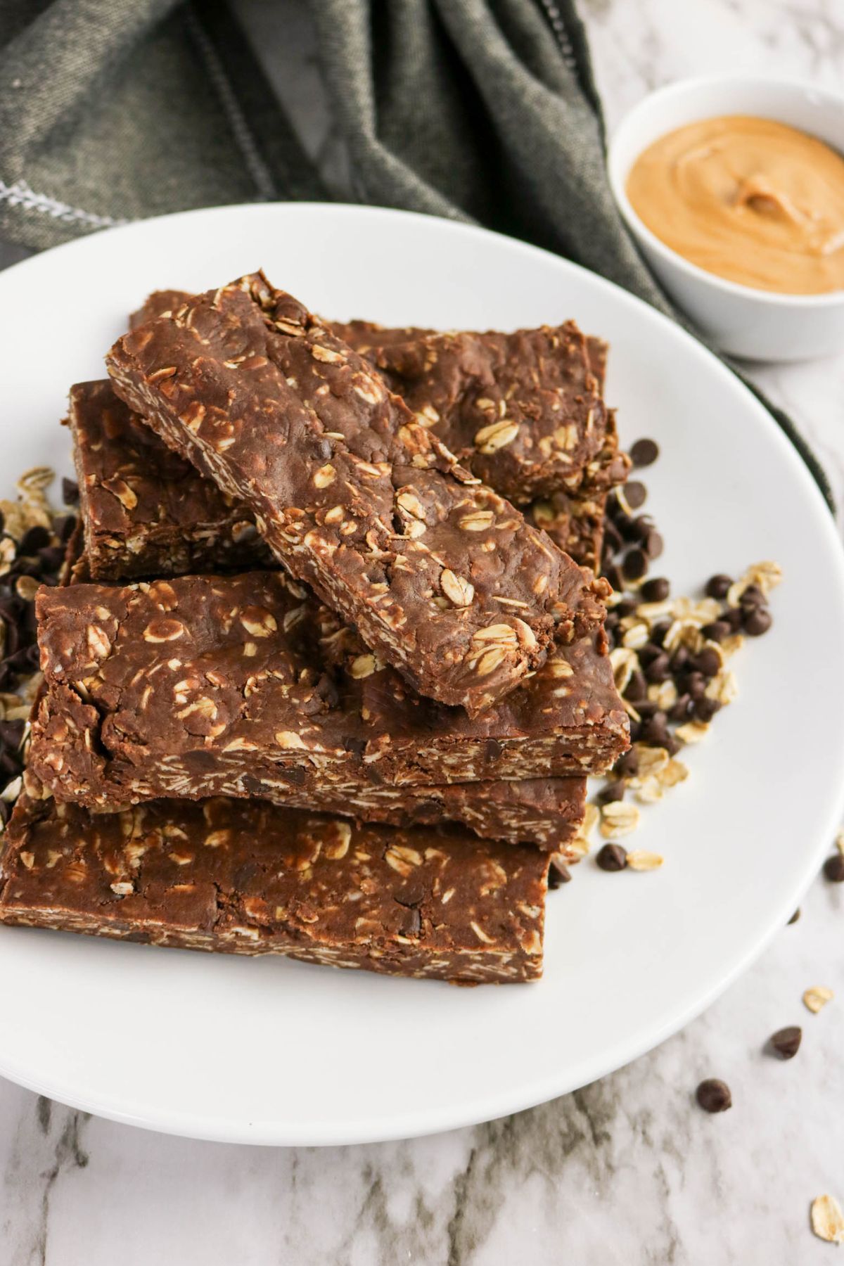 A white plate holds several chocolate oat granola bars stacked on top of each other, surrounded by scattered chocolate chips and oats. A small bowl of peanut butter sits in the background on a marble surface.