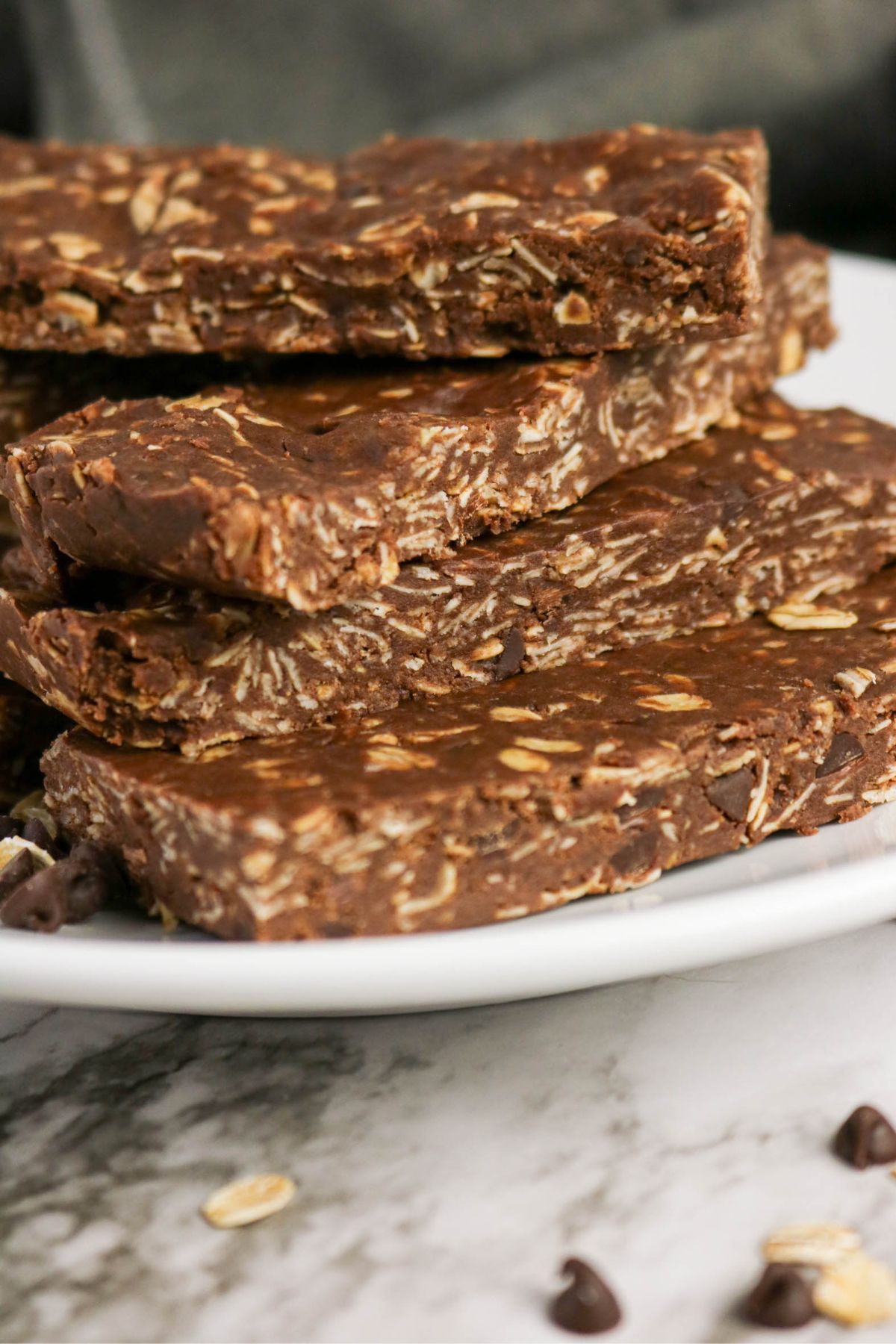 Four chocolate oat bars are stacked on a white plate. The bars have a visible oat texture and are set on a marble surface with a few oats and chocolate chips nearby.