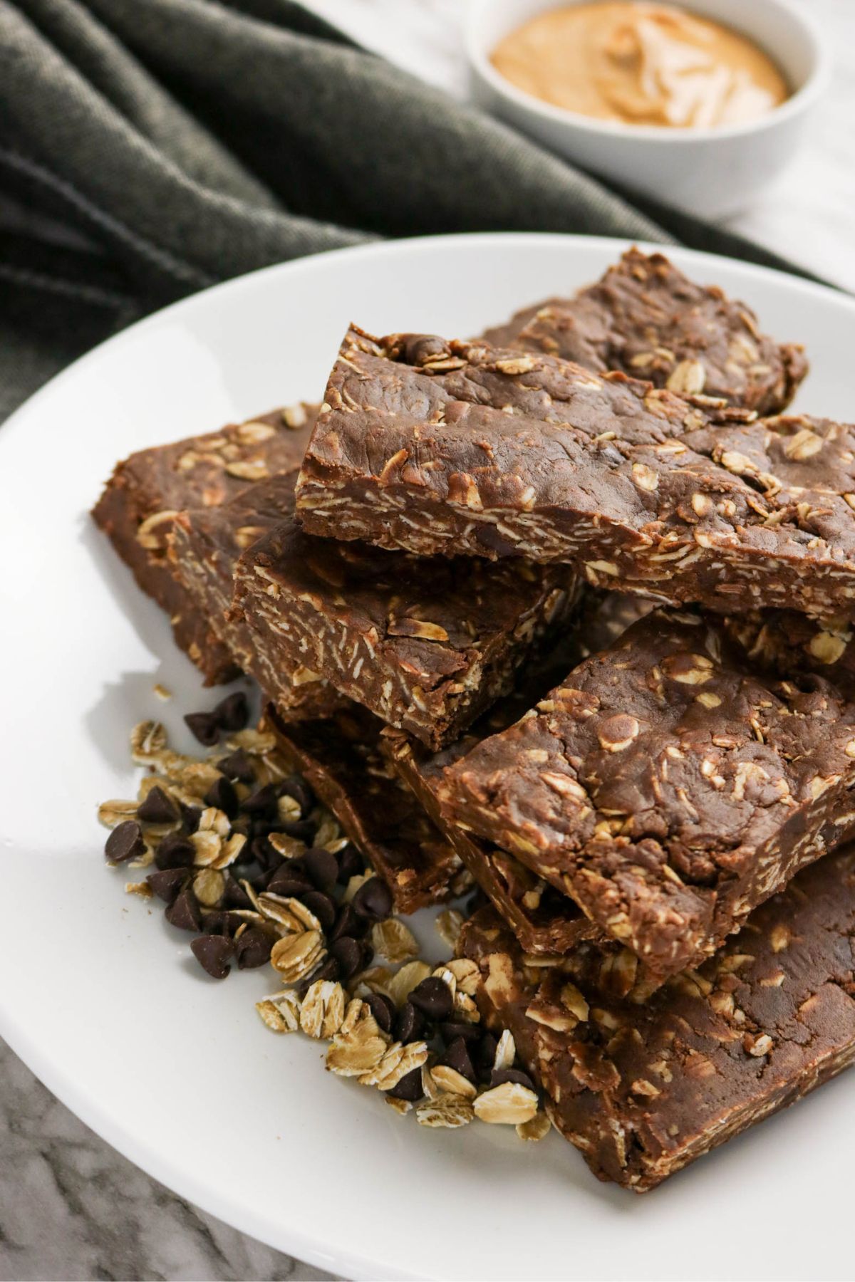 A white plate holds stacked chocolate oat bars with visible oats and chocolate chips, next to small piles of oats and chocolate chips. A bowl of peanut butter and a dark cloth are in the background.