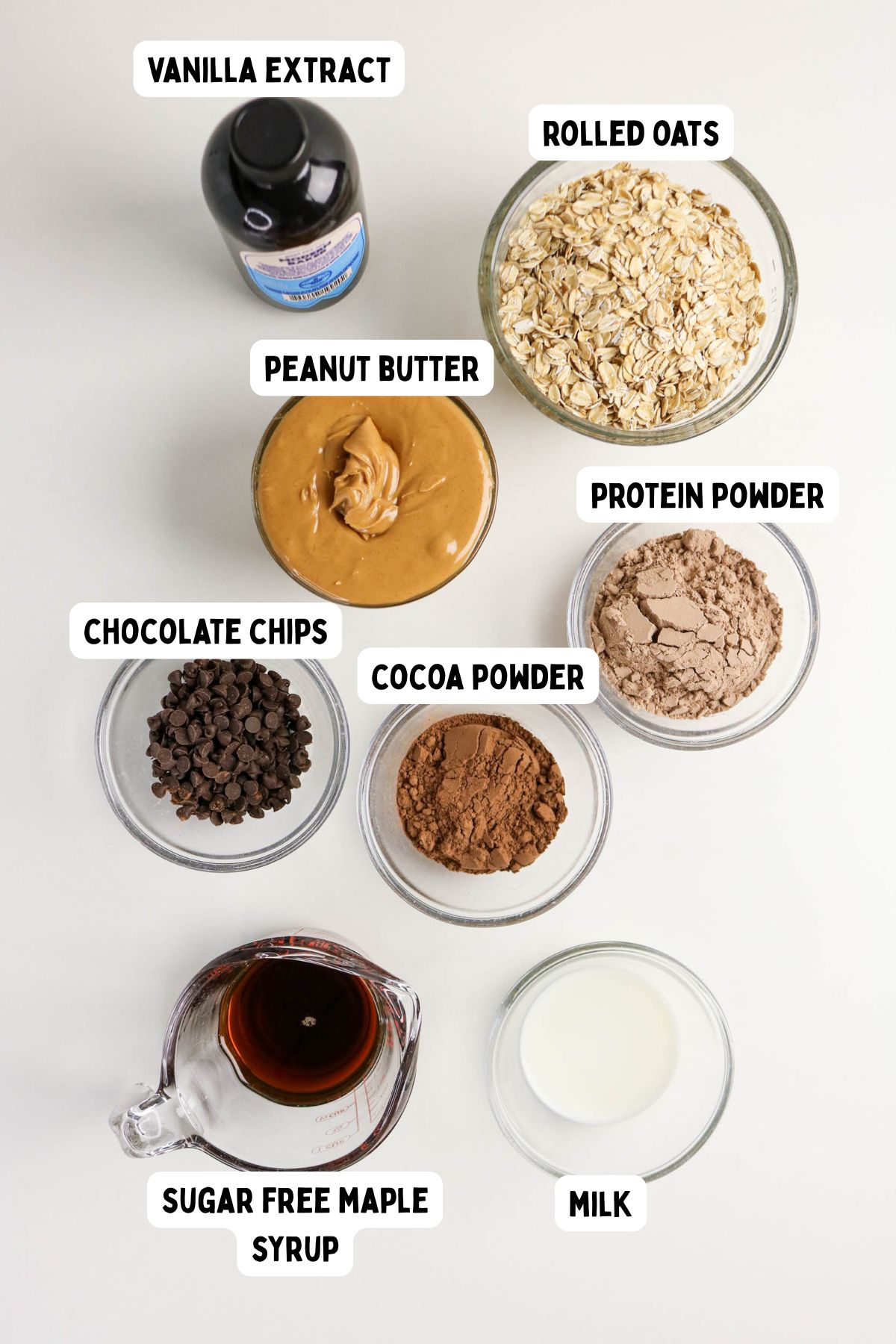 A flat lay of ingredients in bowls and cups labeled as vanilla extract, rolled oats, peanut butter, protein powder, chocolate chips, cocoa powder, sugar free maple syrup, and milk on a white surface.