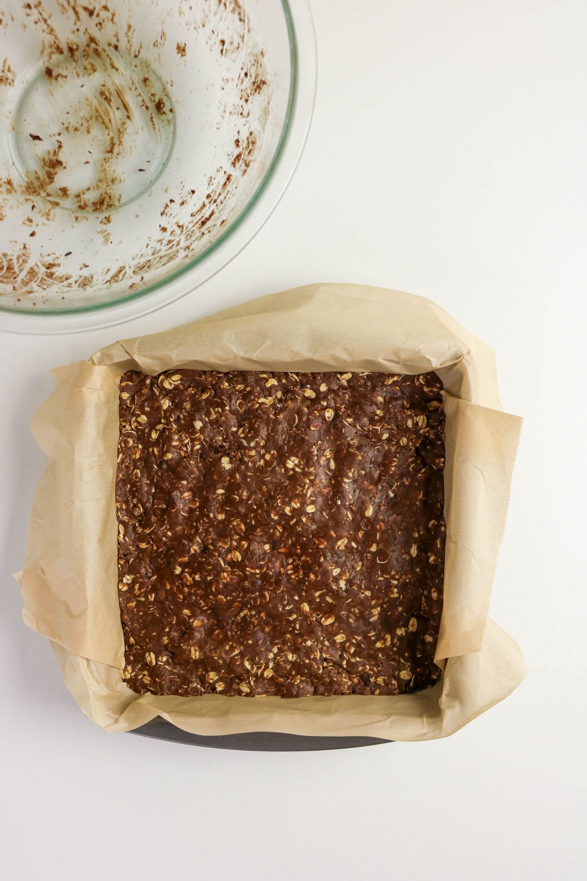 A square baking pan lined with parchment paper holds an unbaked mixture of oats and chocolate. A glass bowl with residue from mixing sits nearby on a white surface.