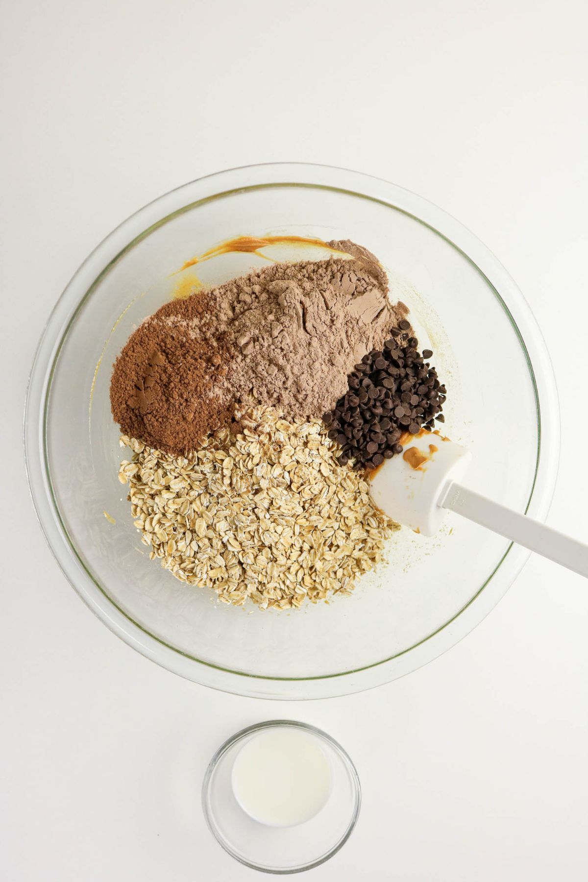 A glass mixing bowl containing cocoa powder, oats, chocolate chips, and peanut butter, with a white spatula inside; a small glass of milk is next to the bowl on a white surface.