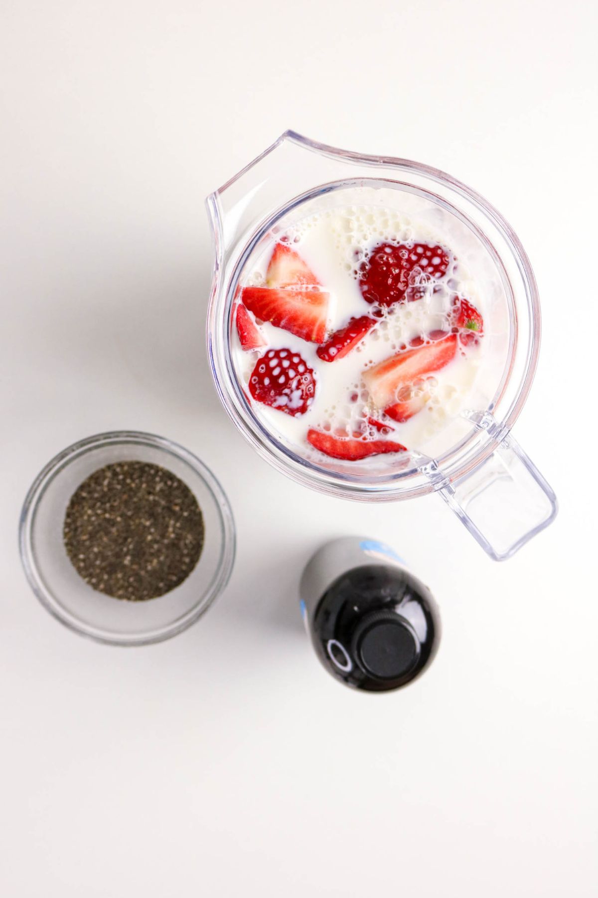 A blender filled with milk, strawberries, and chia seeds sits on a white surface. Next to it are a small glass bowl of chia seeds and a black-capped bottle.