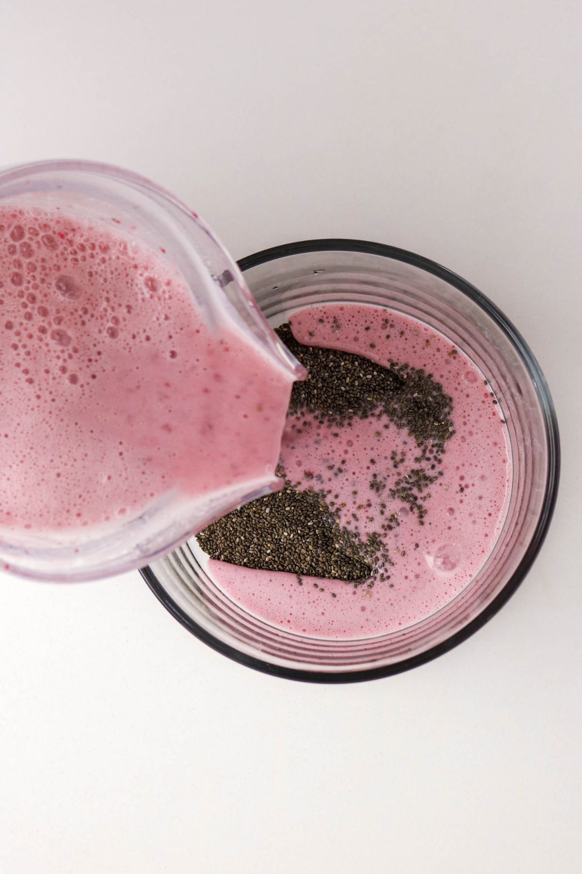 A pink smoothie is being poured from a blender into a glass that contains chia seeds against a white background.